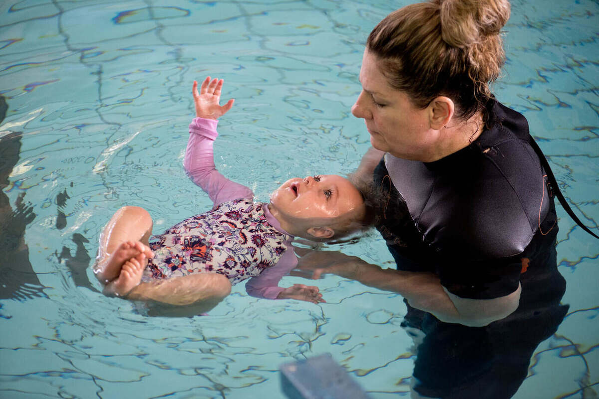 The same infant floating on her back with one arm reaching upward, balanced almost independently while Lizette provides only minimal support beneath the upper back.