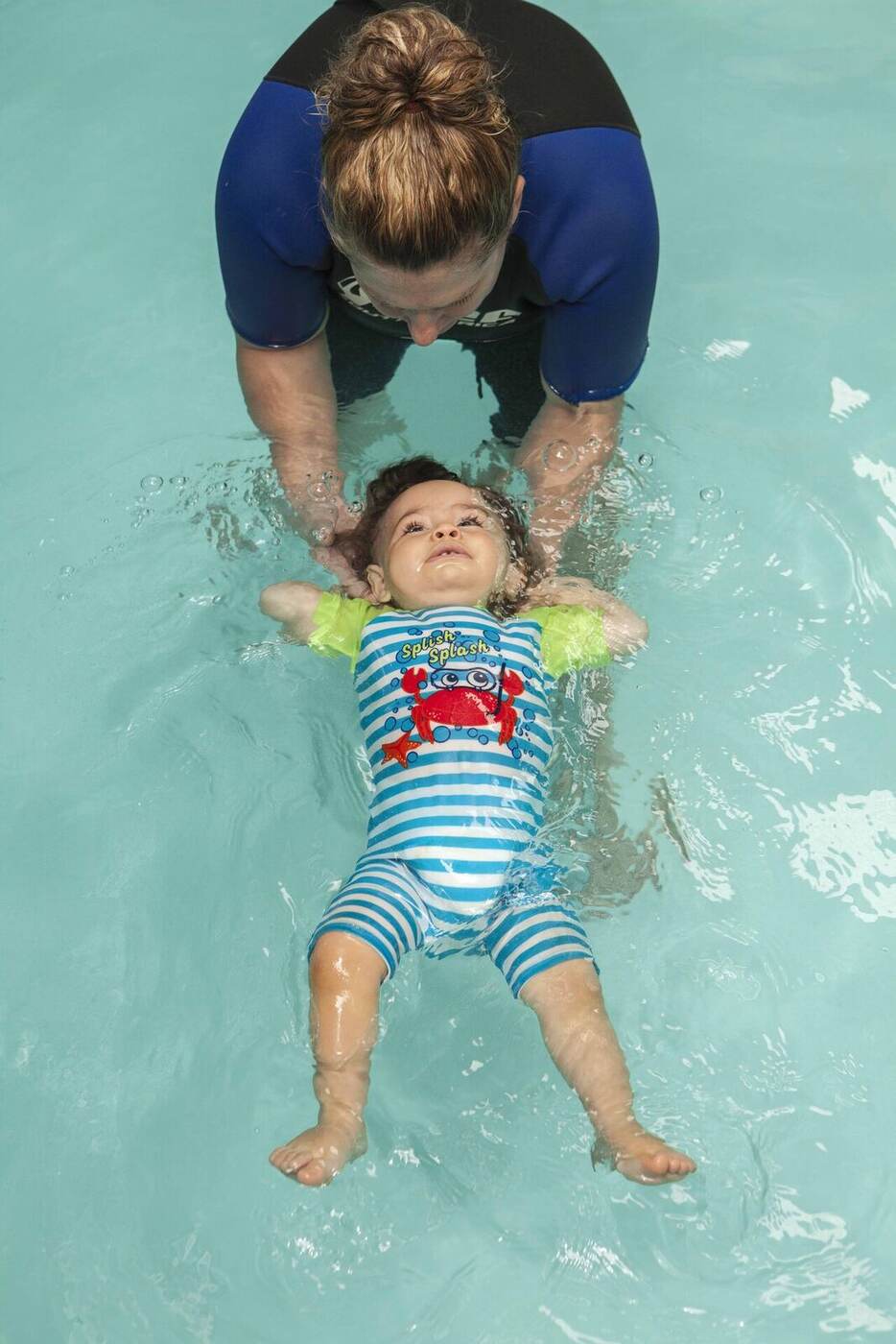 An older baby in a striped Splash Splash romper floating completely unassisted on her back in a starfish position during the Amphibies qualification float, instructor releasing.