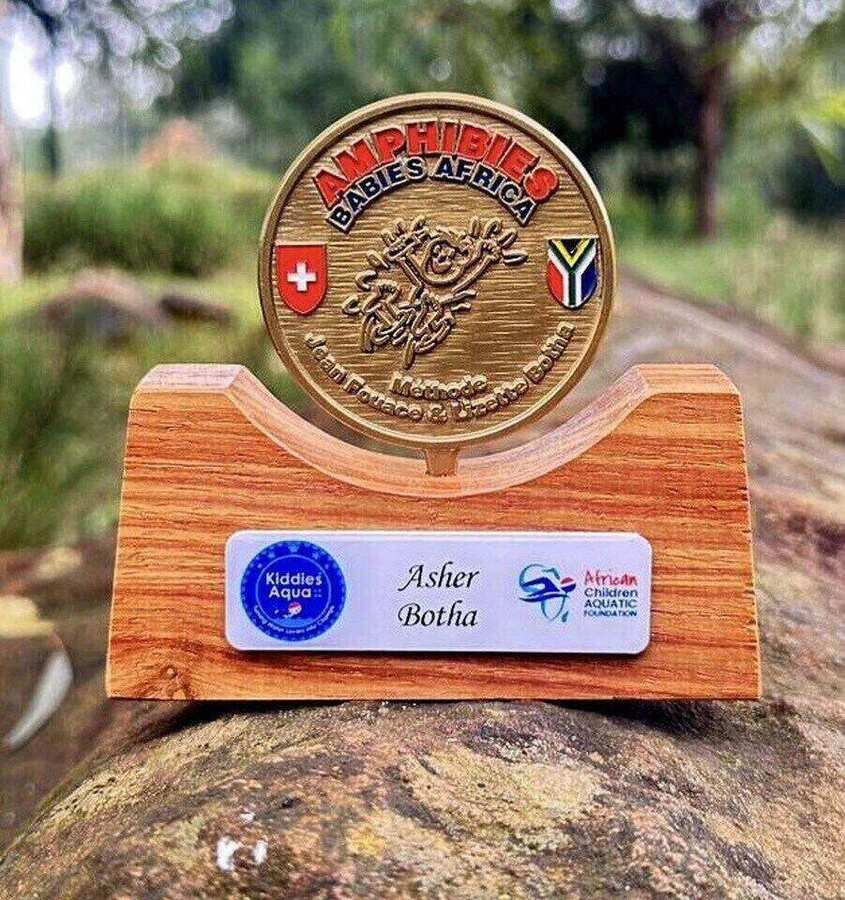 The personalised Amphibies Babies Africa gold-mounted coin awarded to qualifying Kiddies Aqua babies — bronze coin with the Swiss and South African flags, mounted on a wooden stand with the child's name plaque reading 'Asher Botha'.