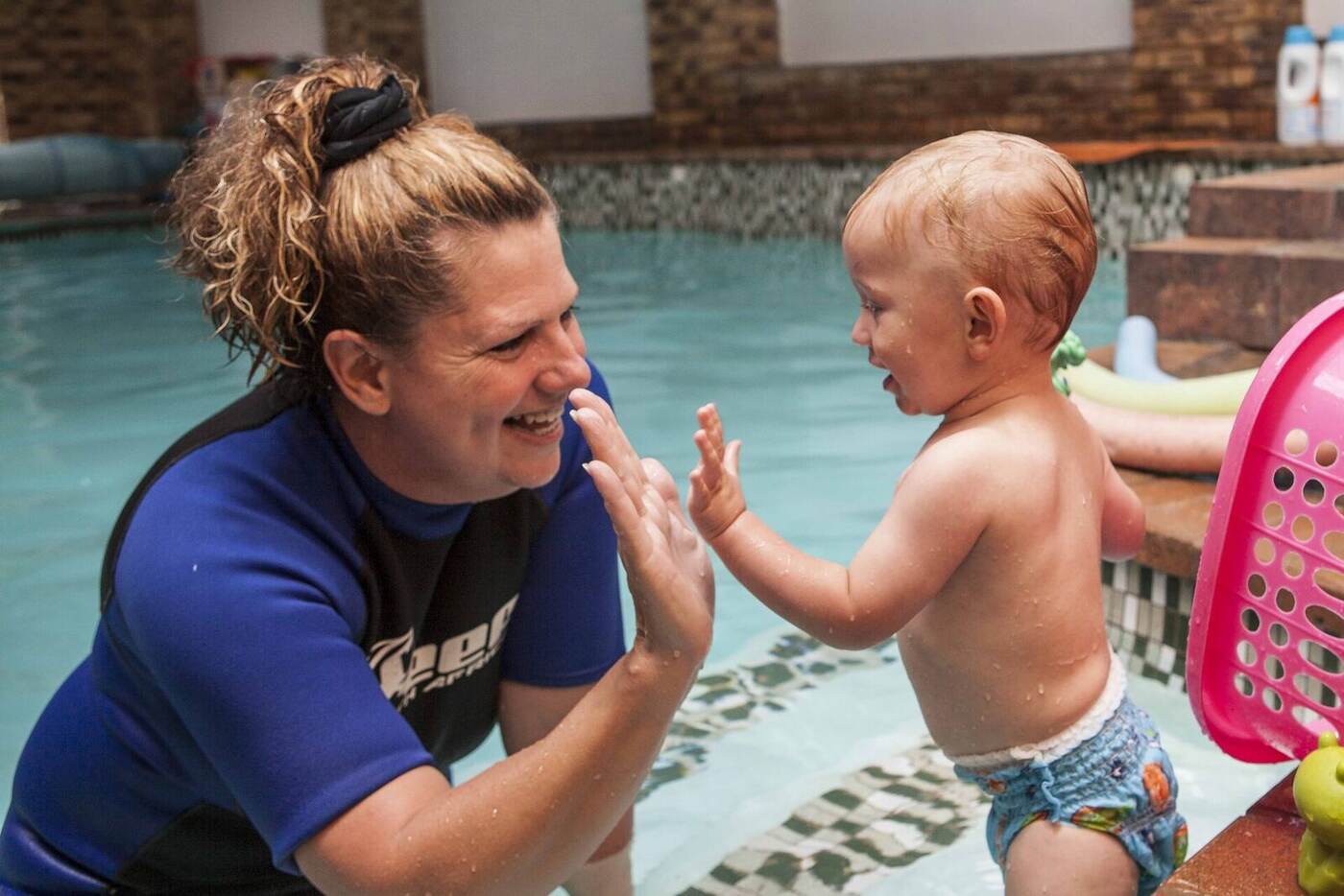 Lizette Botha laughing eye-to-eye with a toddler standing on the pool steps during a water familiarisation lesson at Kiddies Aqua Swimming Academy.