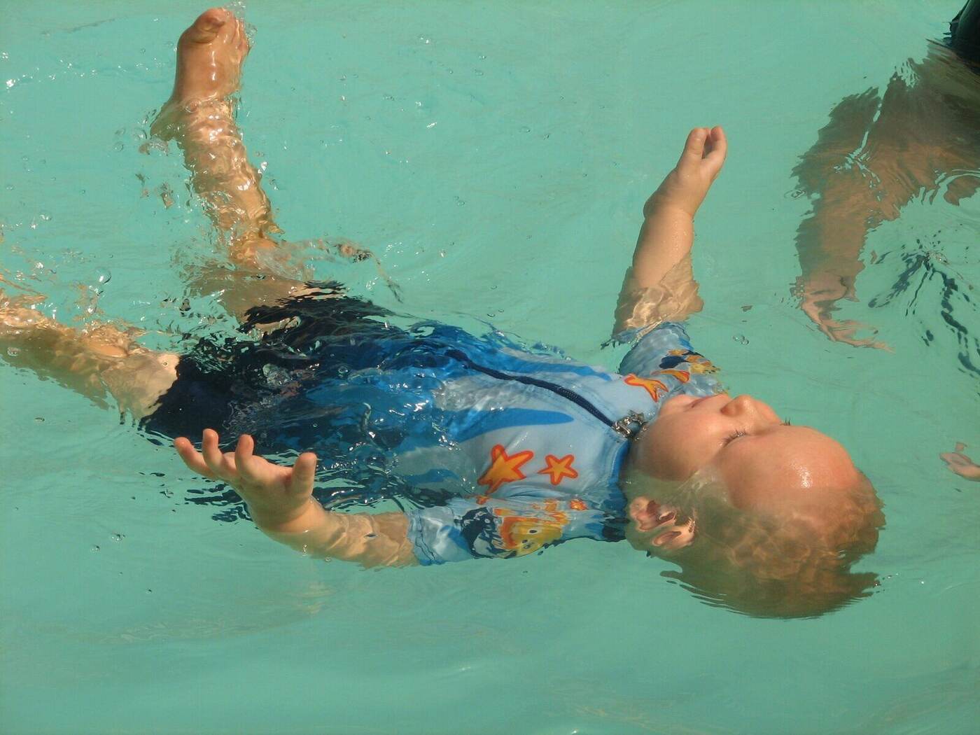 A baby floating fully independently on her back in the pool with no instructor in frame, wearing a long-sleeved patterned costume — the lifelong self-rescue reflex in action.