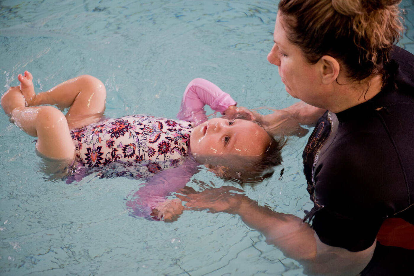 Lizette Botha supporting an infant in a floral costume on her back at the surface of the pool, the baby's head fully cradled in the early supported back-float position.