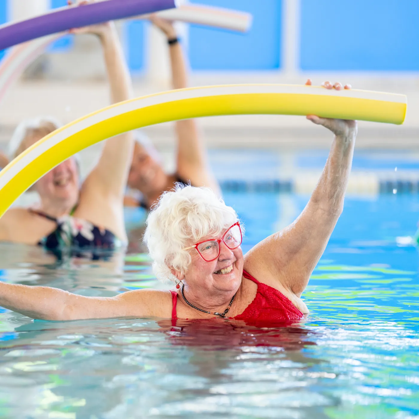 Smiling senior adult raising a pool noodle overhead during a low-impact Kiddies Aqua Water Aerobics class