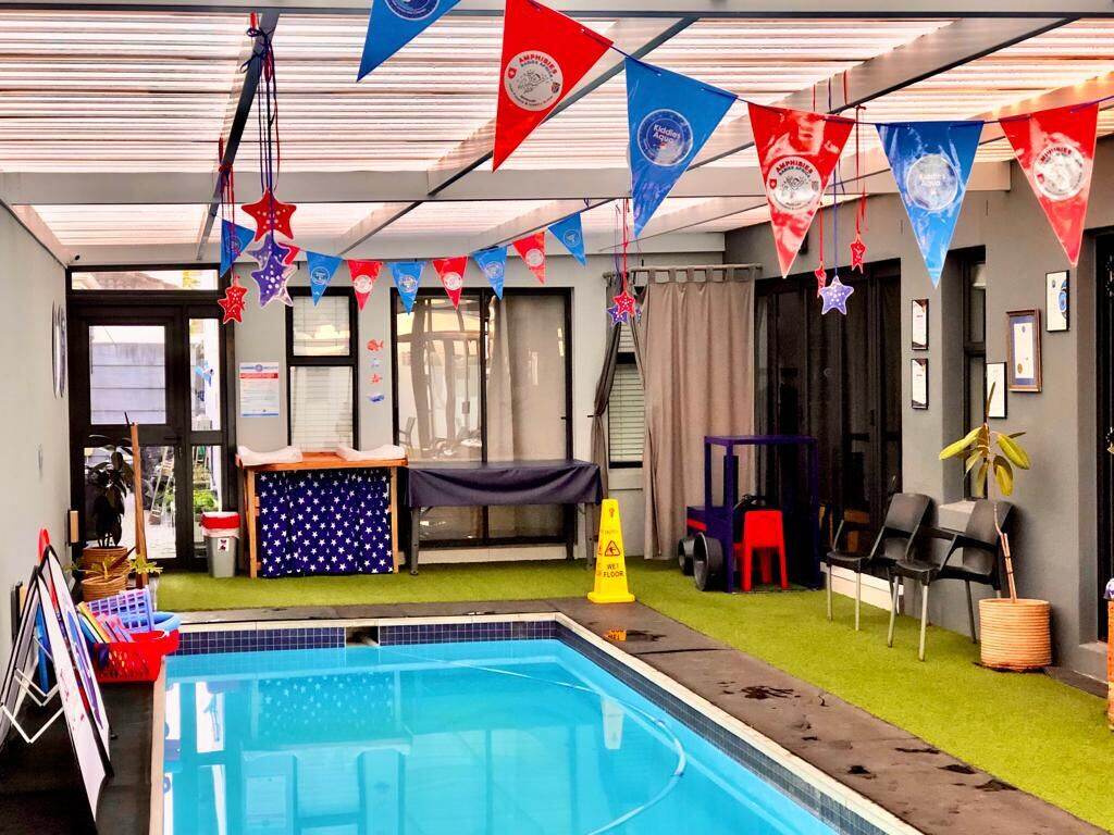 Indoor heated pool dressed with star bunting and branded flags, with artificial turf surrounds and tidy storage for lesson equipment.