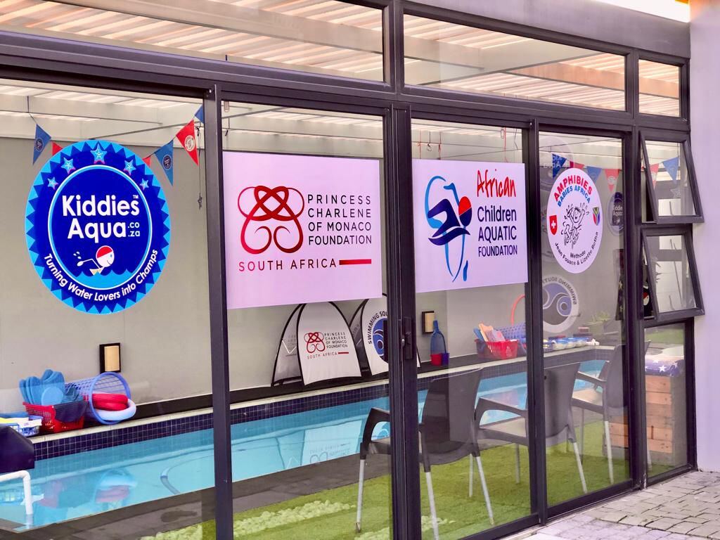 Poolside glass wall displaying the Kiddies Aqua badge alongside the Princess Charlene of Monaco Foundation and African Children Aquatic Foundation partner logos.