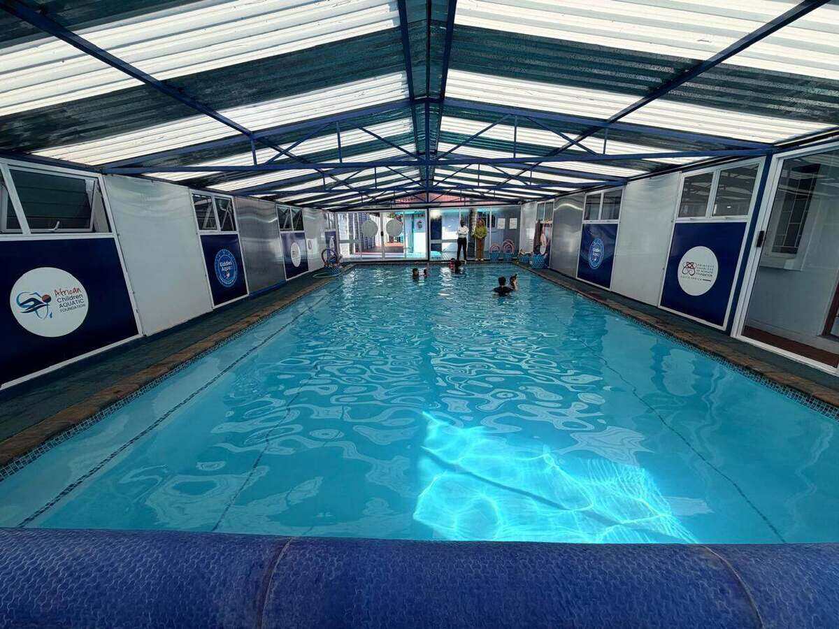 Indoor heated learn-to-swim pool under a covered roof, with calm clear water and partner foundation signage along the poolside walls.