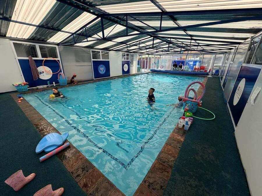 Indoor heated pool under a covered roof with instructors guiding young children through their lessons and equipment laid out on the deck.
