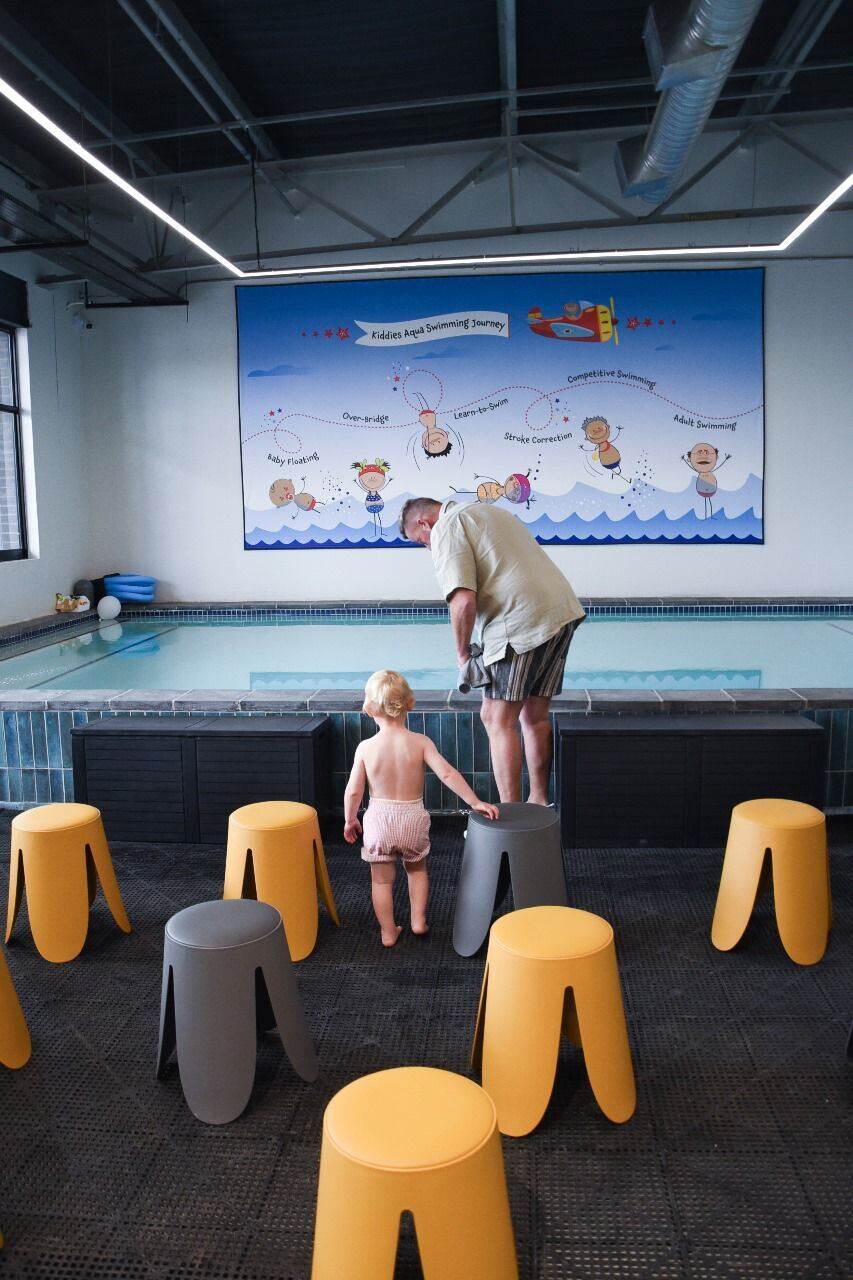 Indoor poolside toddler area featuring the colourful Kiddies Aqua swim-journey mural, with soft seating stools arranged for parents and little ones.