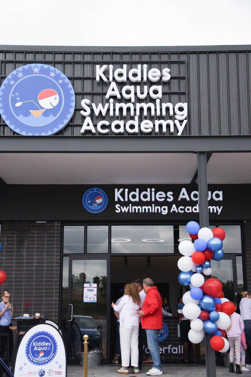 Close-up of illuminated Kiddies Aqua Swimming Academy branch signage above the entrance, framed by red, white and blue grand-opening balloons.