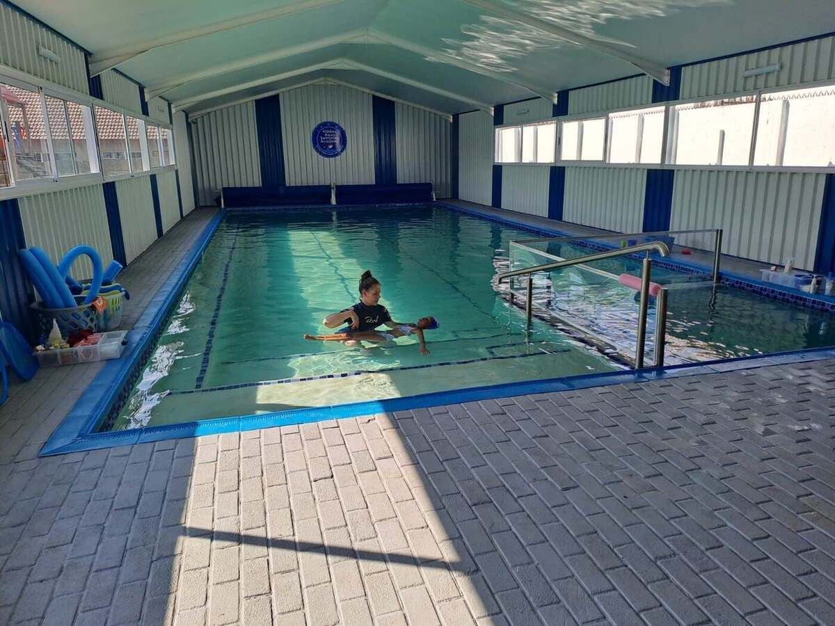An instructor supporting a young swimmer on a kickboard during a lesson in a bright indoor heated pool with a pitched roof.