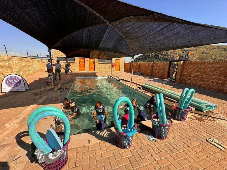 Outdoor learn-to-swim pool shaded by sail nets, with a group lesson in progress and pool noodles in baskets, set against a mountain backdrop.