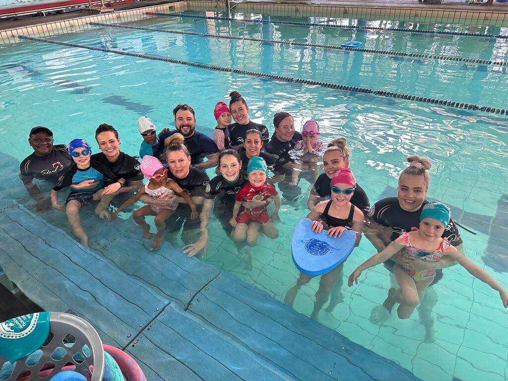 A group of young swimmers and their instructors smiling together at the edge of an indoor lane pool after a lesson.