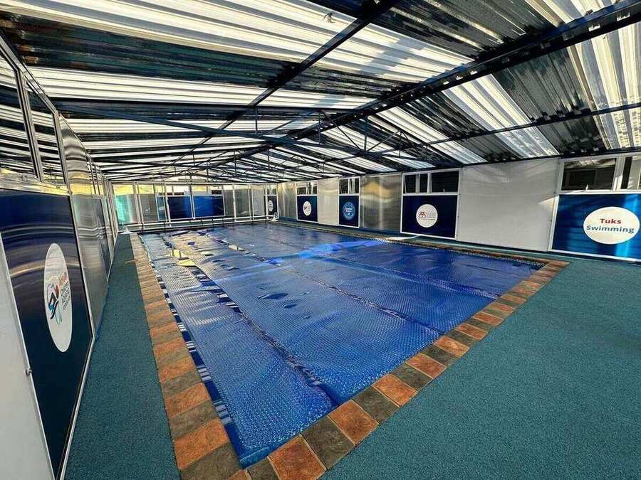 Covered indoor heated pool fitted with a thermal cover, lined with branded partner banners under a clear-span roof.