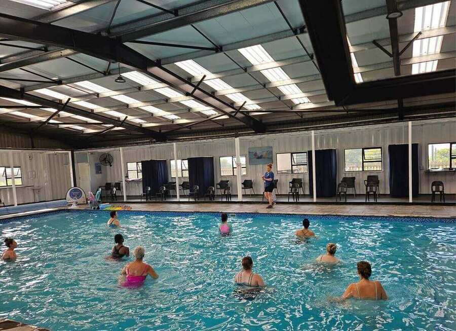 Adults taking part in an aqua-aerobics class in a large covered indoor pool, with an instructor leading from the poolside.